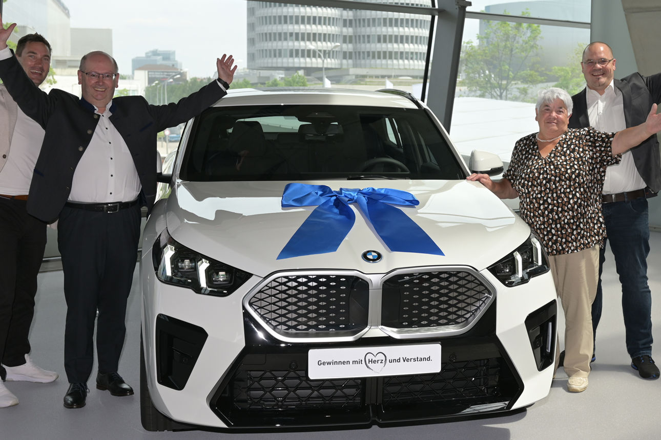 Vier jubelnde Personen mit in die Höhe gereckten Händen stehen um  neuen weißen BMW mit blauer Schleife