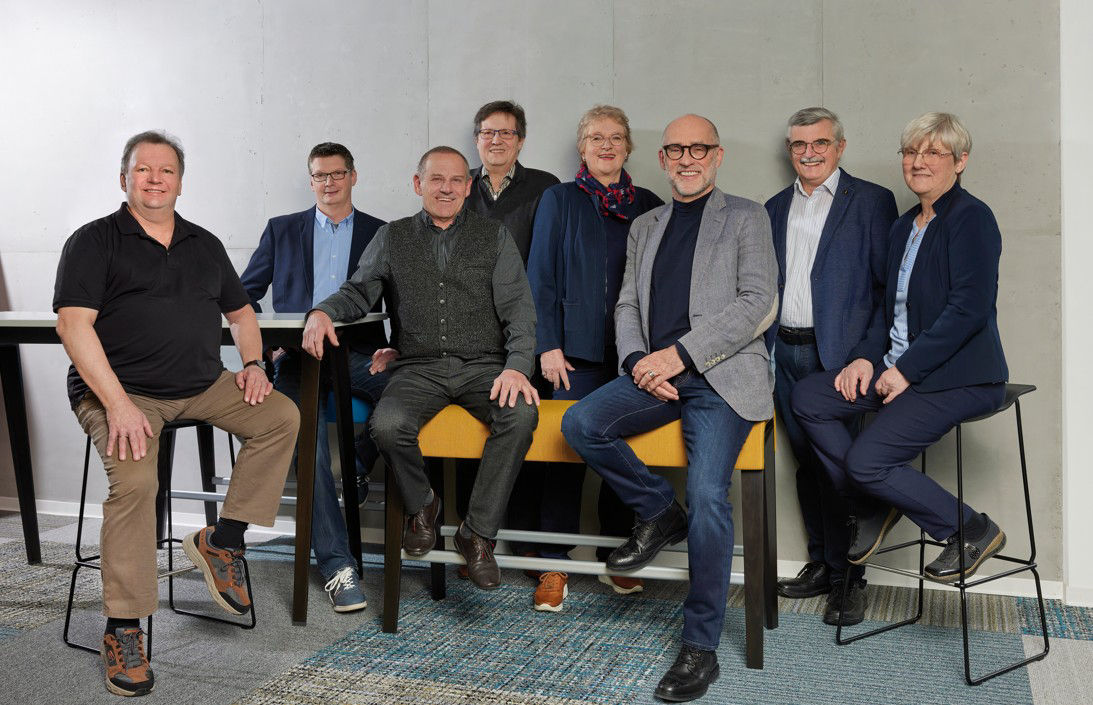 Eine Gruppe von acht Personen, meist Männer, steht in lässiger Pose vor einer hellgrauen Wand.