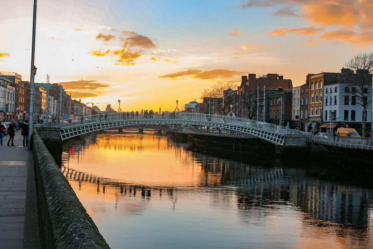 Eine Person geht links am Ufer der Liffey in Dublin entlang. Mittig quert die belebte Ha'penny Bridge den Fluss, dahinter die