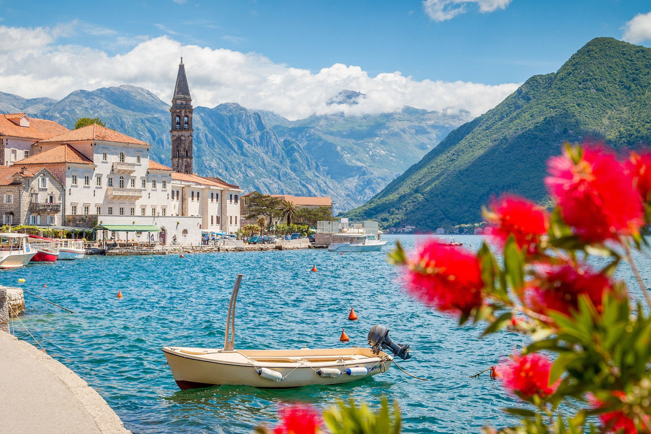 Ein kleines Boot treibt vor Perast im türkisblauen Wasser der Bucht von Kotor in Montenegro. Am Ufer stehen historische Stein