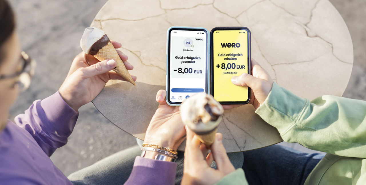 Zwei Personen sitzen mit einem Eis an einem Bistrotisch. Eine Person schickt der anderen Person Geld mit Wero.