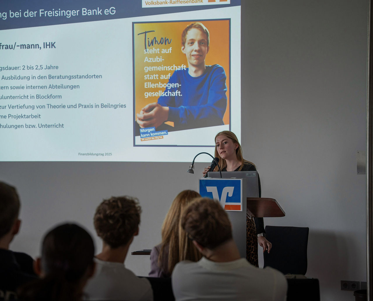 Emma Kellerer steht hinter Rednerpult, hinter ihr ein Teil ihrer Präsentation und spricht zu Schüler/Innen, die von hinten zu sehen sind. 