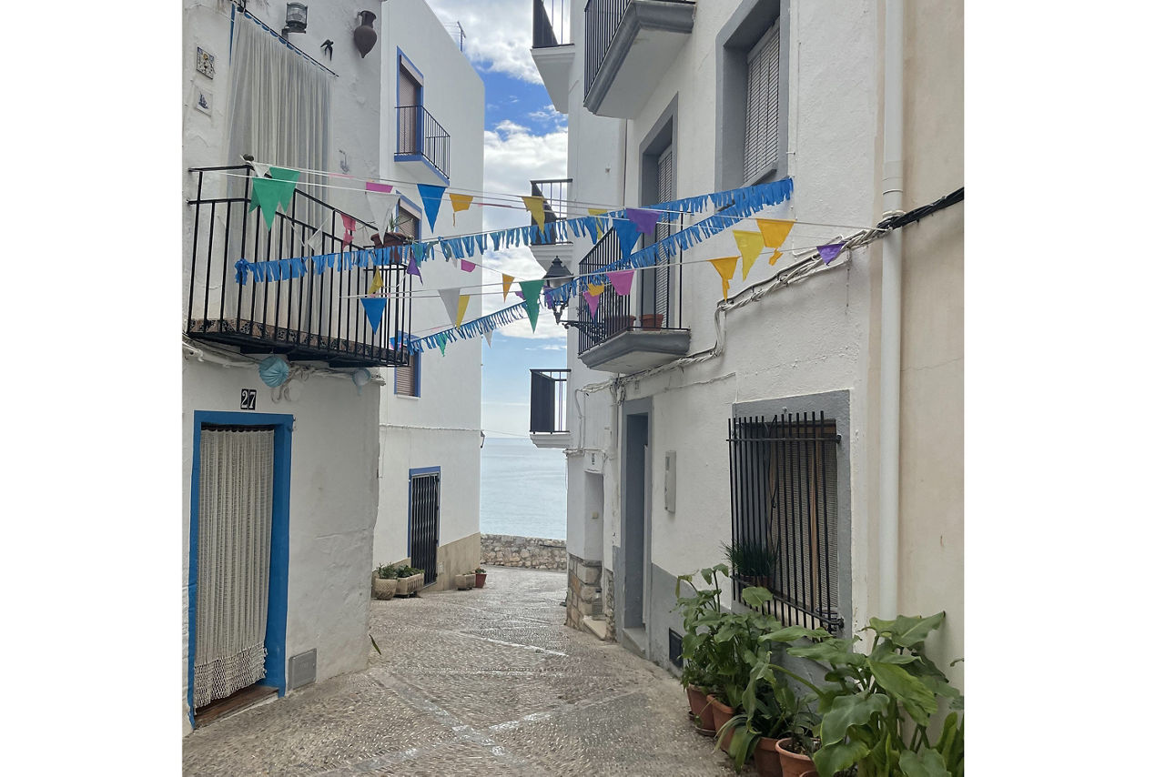Schmale Gasse mit weiß getünchten Häusern, bunten Wimpelketten und Blumentöpfen, Blick auf das Meer am Ende der Straße.