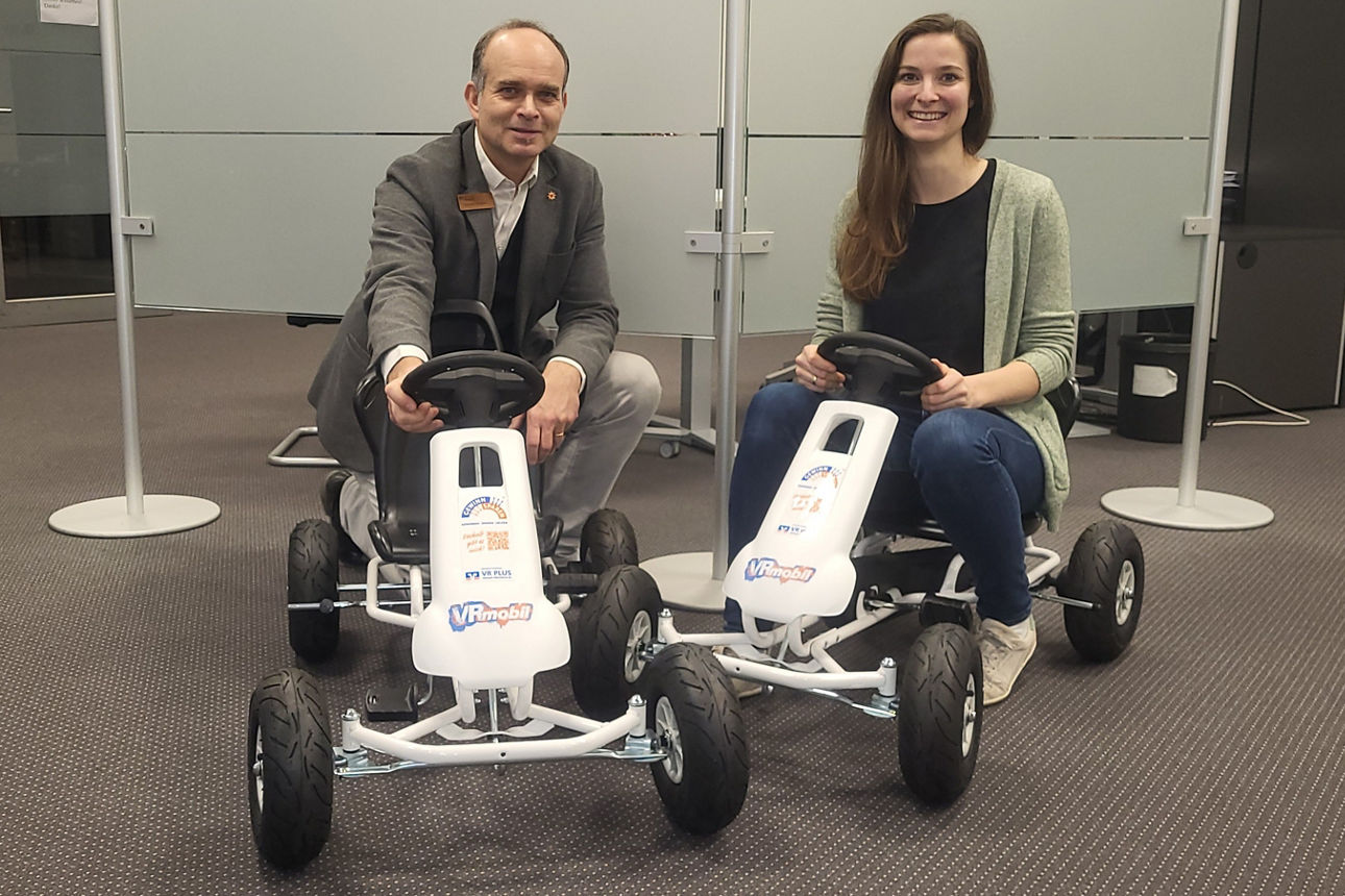 Zwei Bankmitarbeiter sitzen auf zwei Gokarts im Eingangsbereich der Bank. 