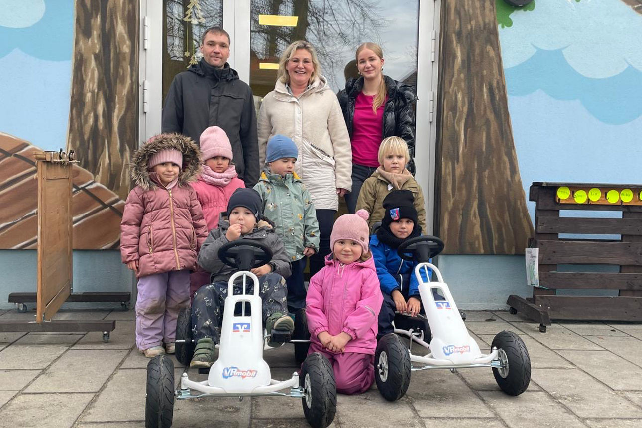 Drei Erwachsene und sieben Kita-Kinder stehen vor dem Kita-Eingang mit zwei Gokarts.
