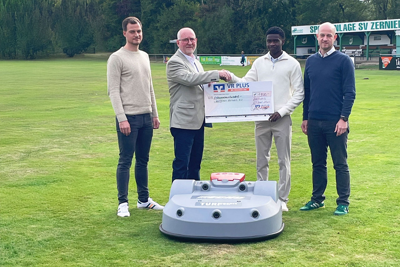 vier Männer stehen auf einem Rasen und halten einen Scheck hoch, vor ihnen steht ein großer Mähroboter
