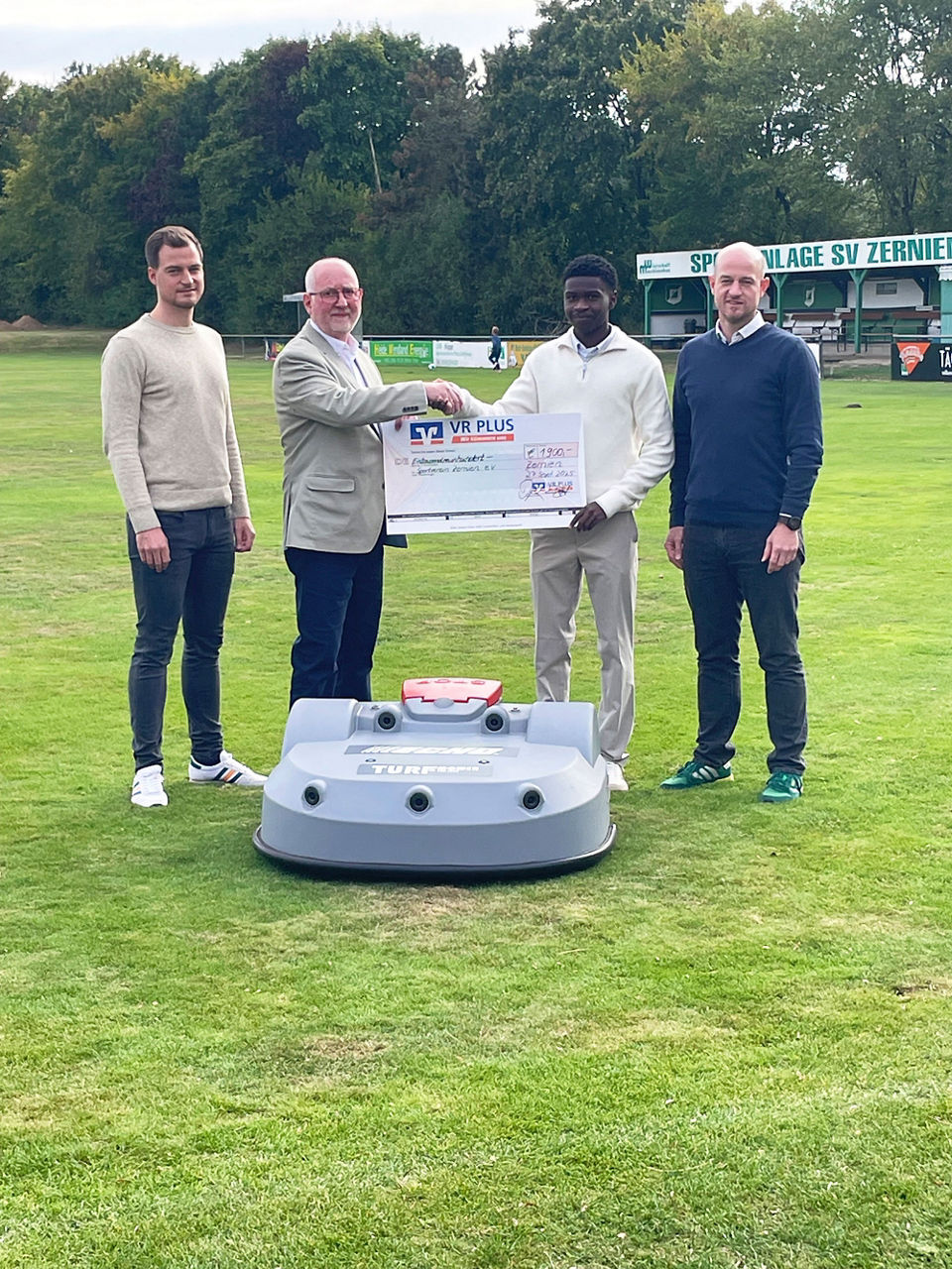 vier Männer stehen auf einem Rasen und halten einen Scheck hoch, vor ihnen steht ein großer Mähroboter