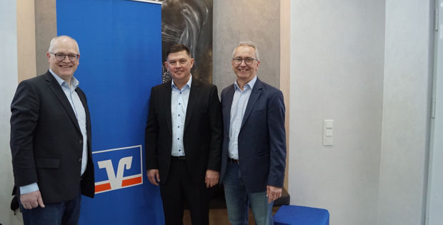 Drei Männer stehen vor einem blauen Banner mit einem weißen „V“-Logo.