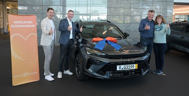 Zwischen 4 Personen steht ein dunkelgraues Auto mit einer blau-orangen Schleife auf der Motorhaube