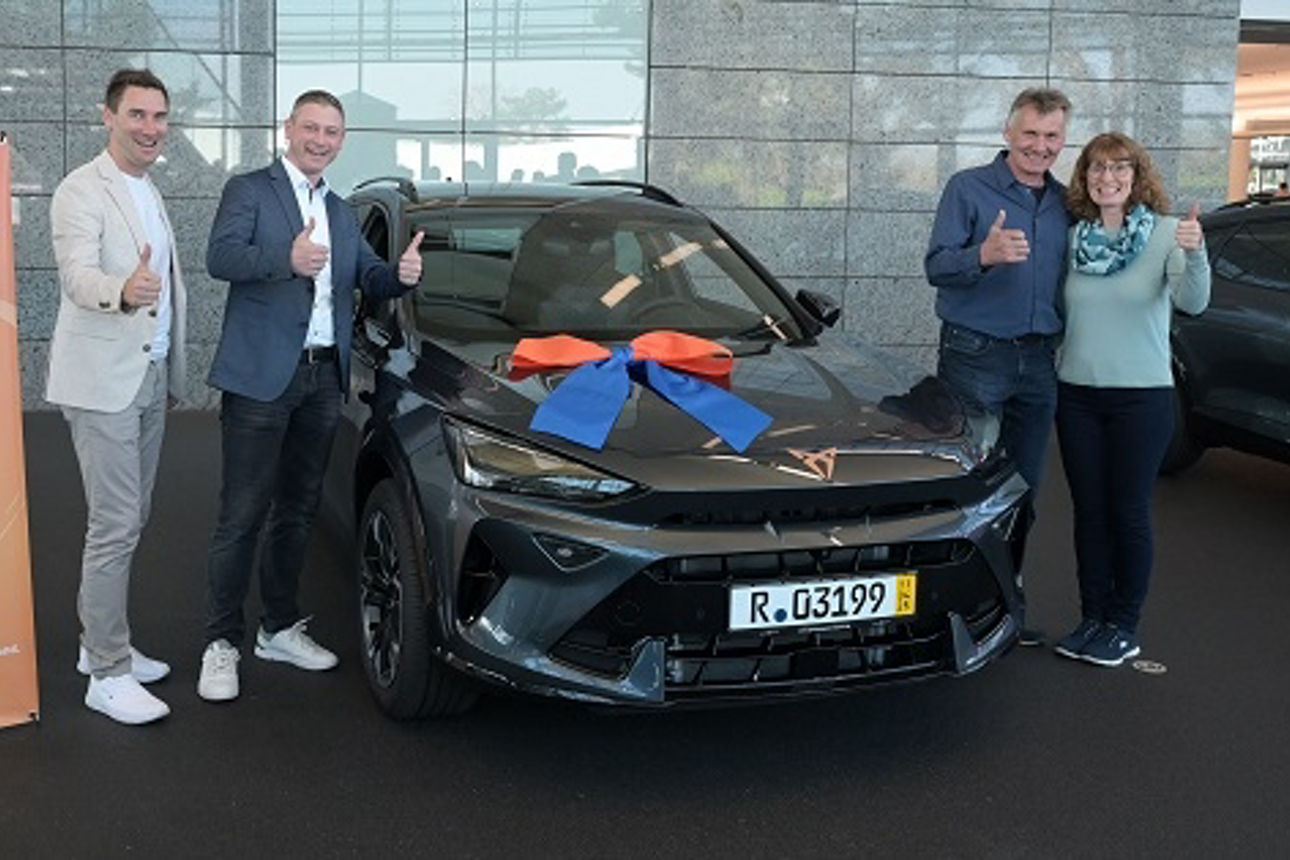 Zwischen 4 Personen steht ein dunkelgraues Auto mit einer blau-orangen Schleife auf der Motorhaube