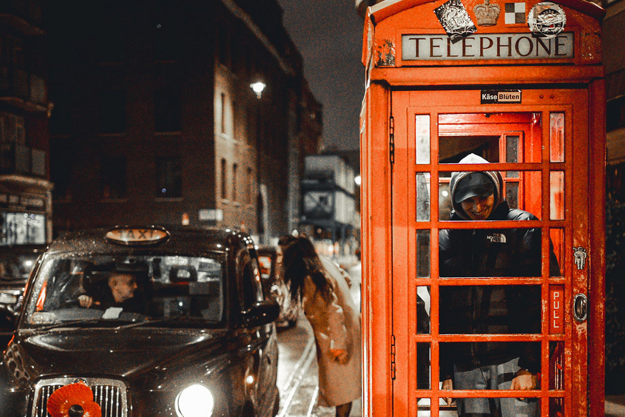 Rote Telefonzelle mit Person darin, daneben schwarzes Taxi mit rotem Mohnblumen-Aufkleber auf der Motorhaube, nächtliche Straßenszene in London.
