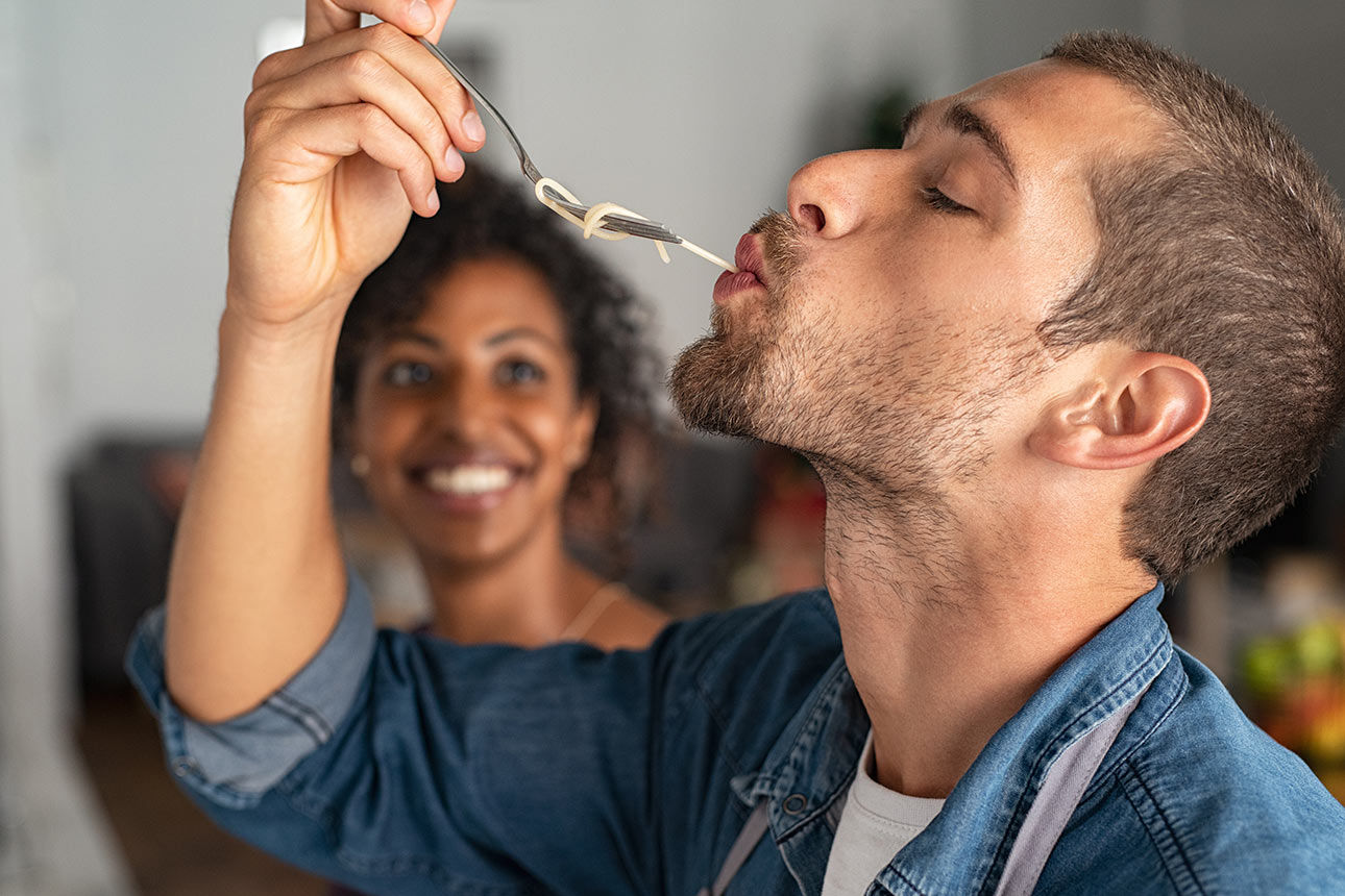 Mann der eine Spagetti von einer Gabel schlürft. Eine Frau im Hintergrund