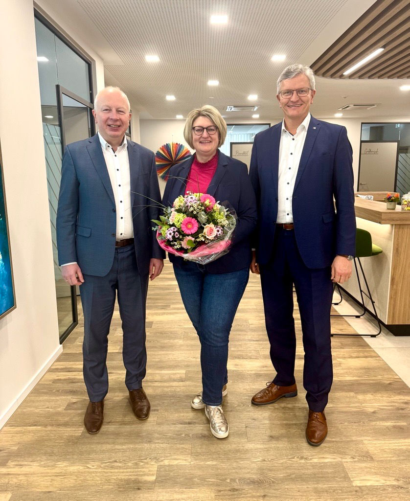 zwei Männer und eine Frau mit bunten Blumenstrauß stehen nebeneinander und lächeln in die Kamera