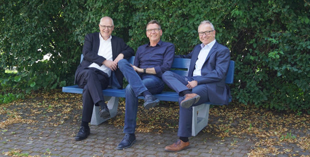 Drei Männer in Business-Kleidung sitzen auf einer blauen Bank.