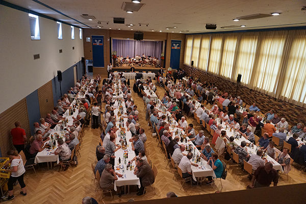Das Bild zeigt einen großen Saal, der mit Menschen gefüllt ist, die an langen Tischen sitzen.