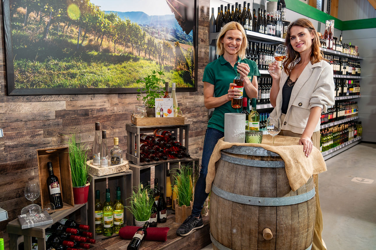 Zwei Frauen stehen an einem Weinfass und stoßen mit zwei Weingläsern an.