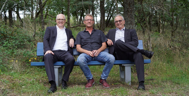 Drei Männer sitzen auf einer blauen Bank in einem bewaldeten Gebiet.