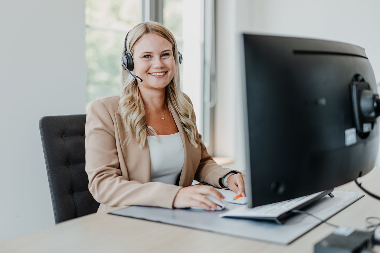 Frau mit Headset sitzt lächelnd am Schreibtisch vor einem Bildschirm