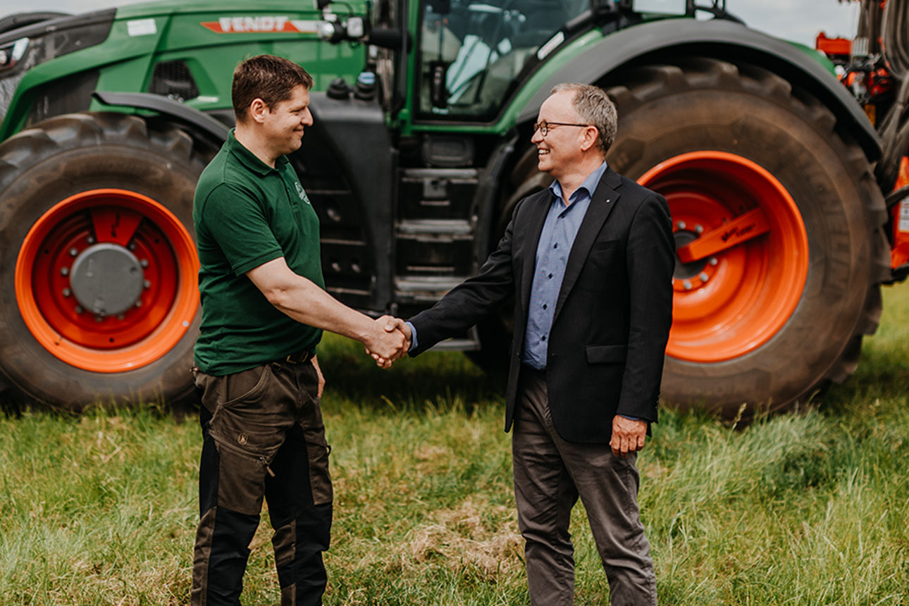 Zwei Männer stehen in Business Kleidung auf einem Feld vor einem Traktor.