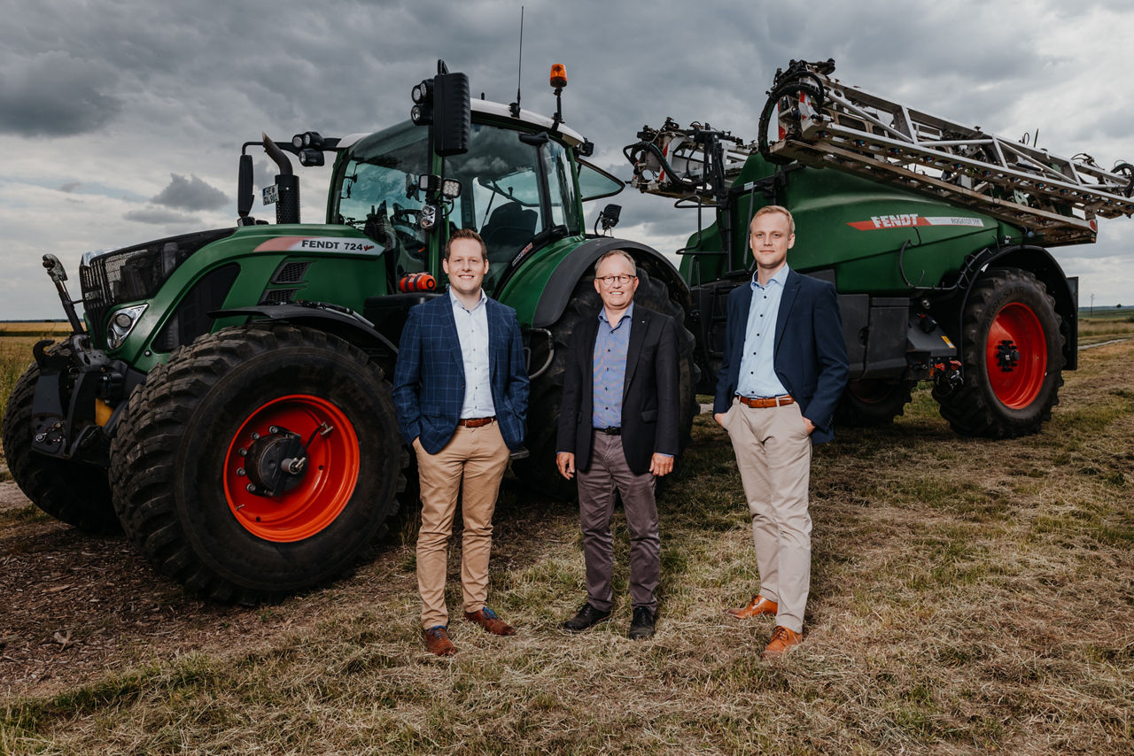 Drei Männer in Business Kleidung stehen auf einem Feld vor einem Traktor.