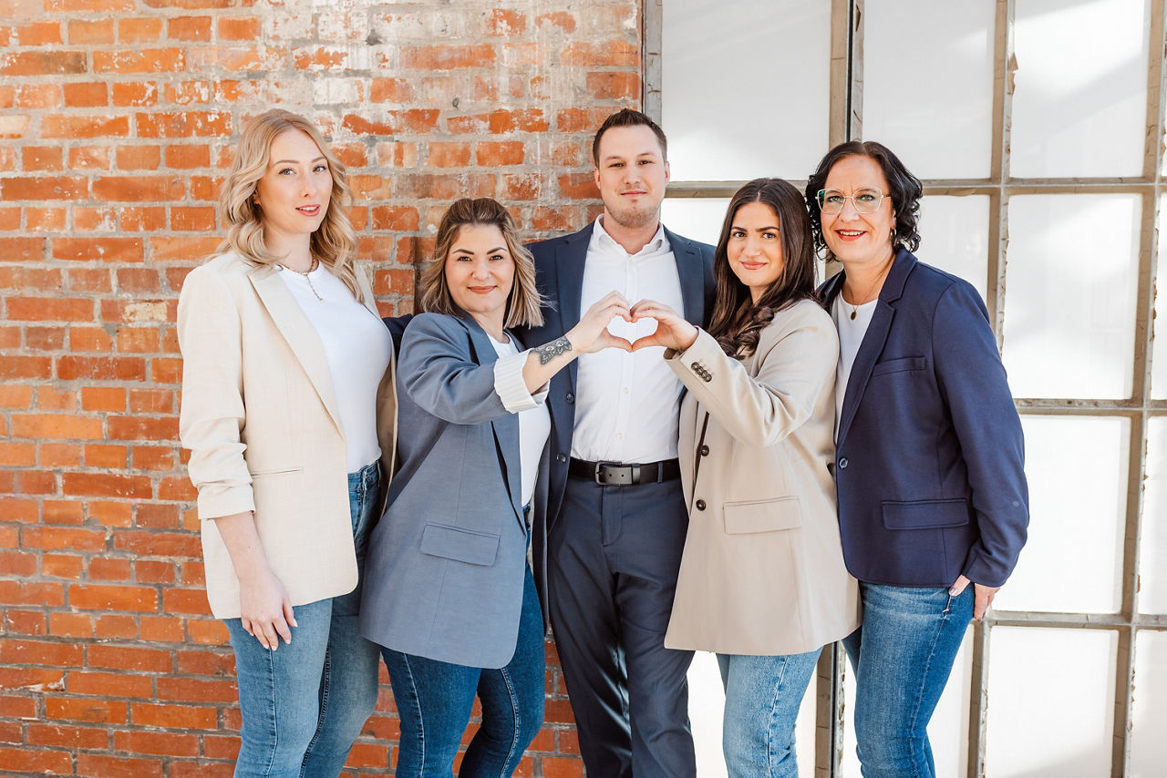 Fünf Personen stehen zusammen für ein Gruppenbild und zwei Frauen formen mit jeweils einer Hand zusammen ein Herz