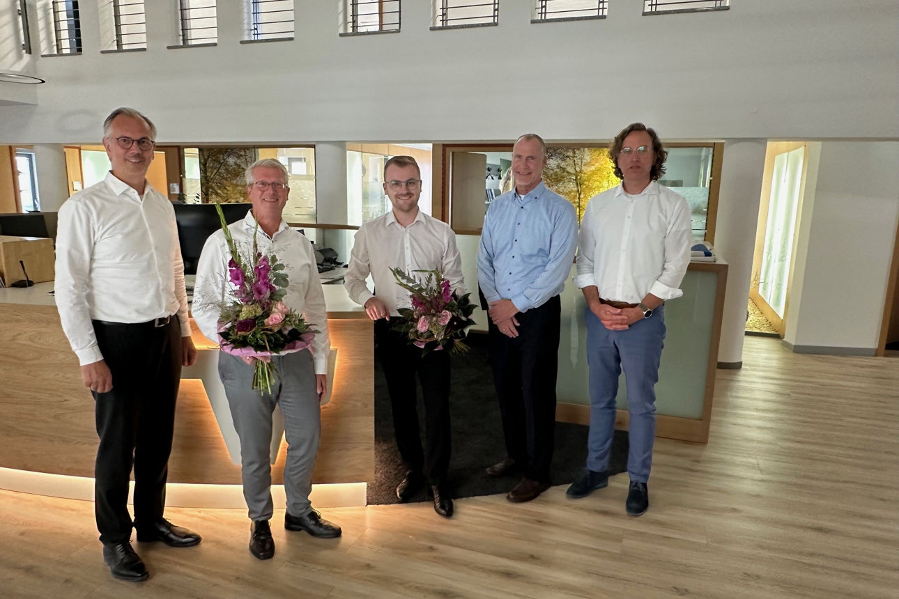 Fünf Männer in formeller Kleidung stehen mit Blumensträußen in einem modernen Büro.