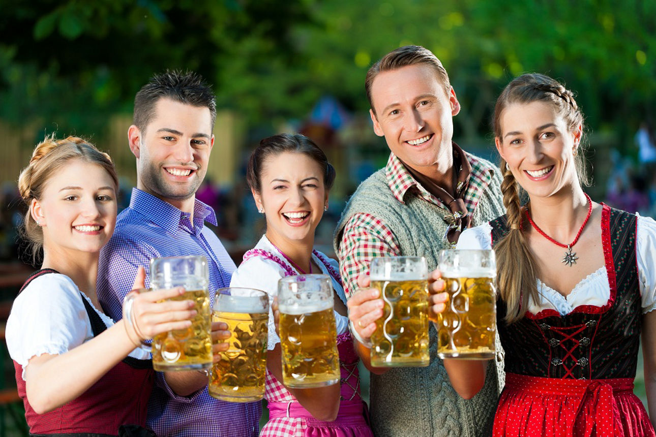 Personen stehen im Freien vor einem unscharfen Hintergrund mit grünen Bäumen und halten gefüllte Bierkrüge in die Kamera.