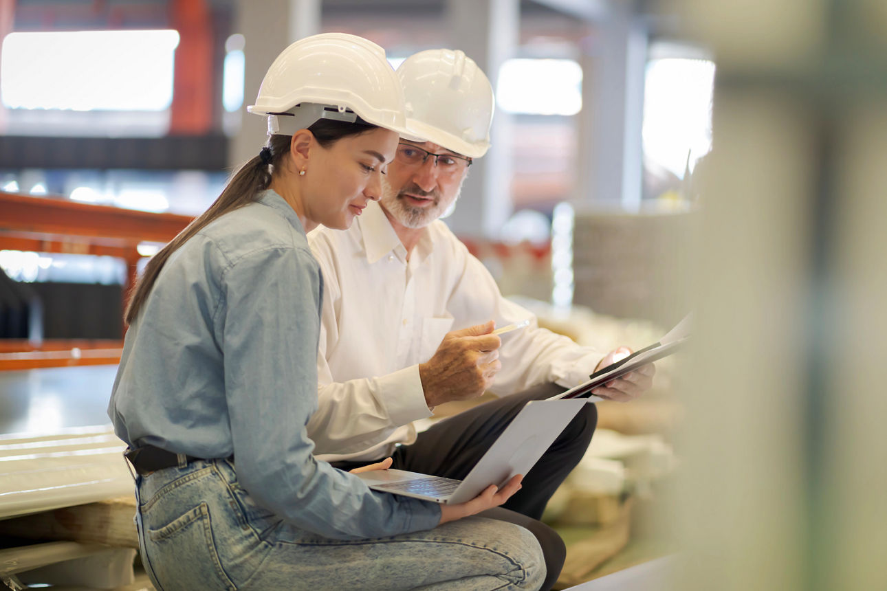 Mann und Frau mit Schutzhelm auf dem Kopf sitzen zusammen und schauen gemeinsam auf den Laptop der Frau