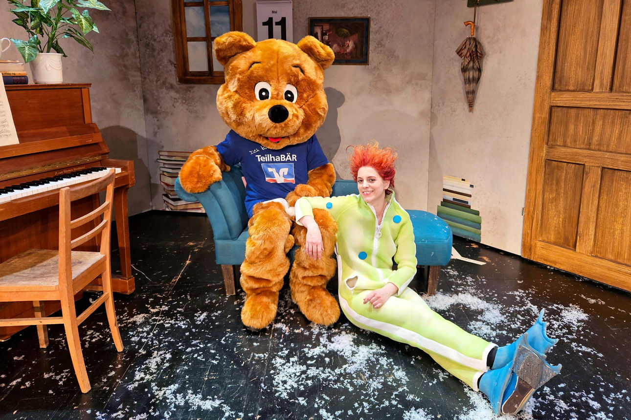 Maskottchen der VR-Bank Mittelsachsen sitzt neben der Schauspielerin im Bühnenbild