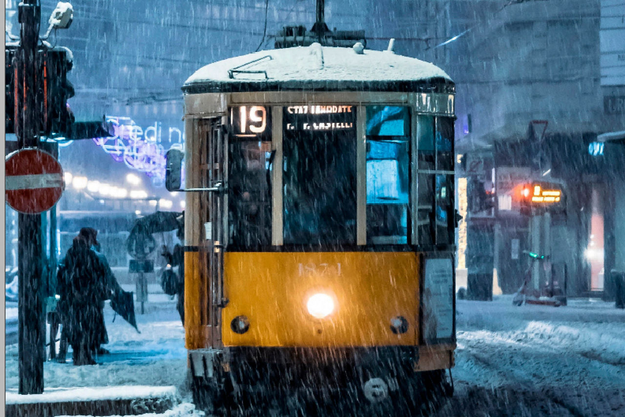 Gelber Straßenbahnwagen der Linie 19 bei Schneefall in der Nacht, umgeben von Fußgängern mit Regenschirmen.