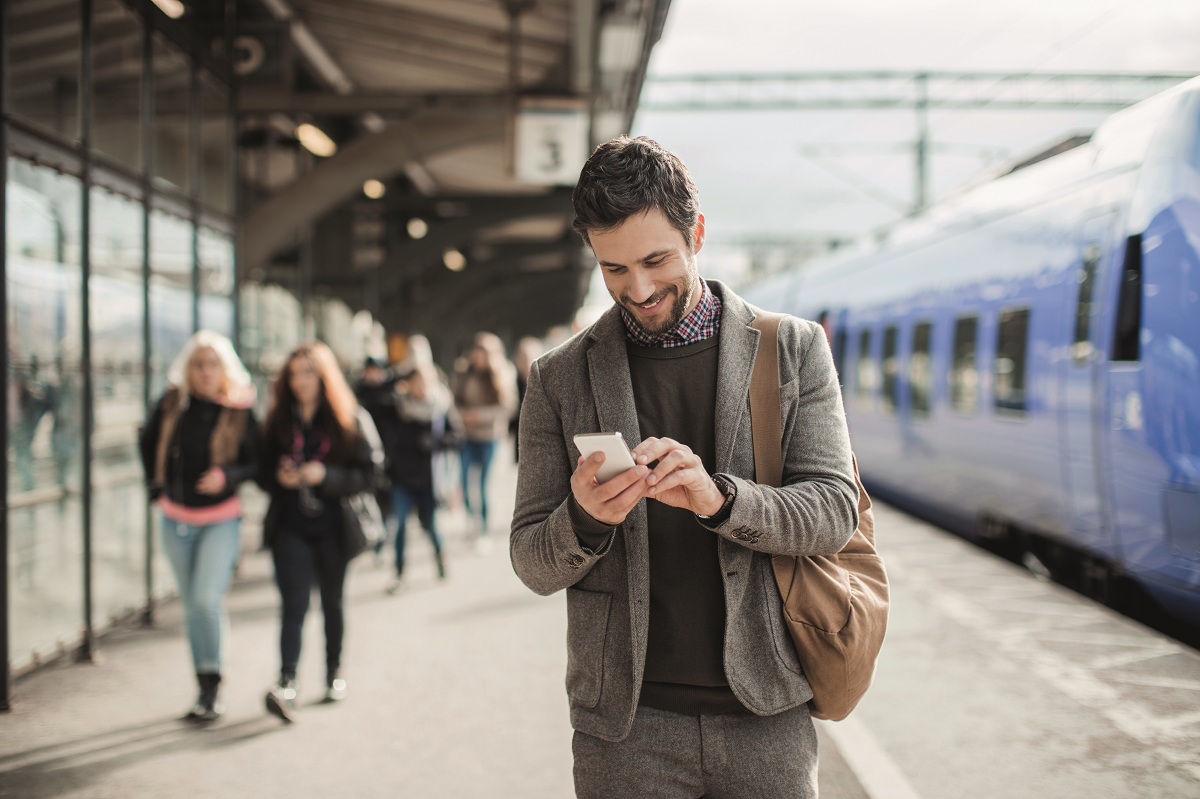 Mann steht an Bahnsteig und tippt auf Smartphone