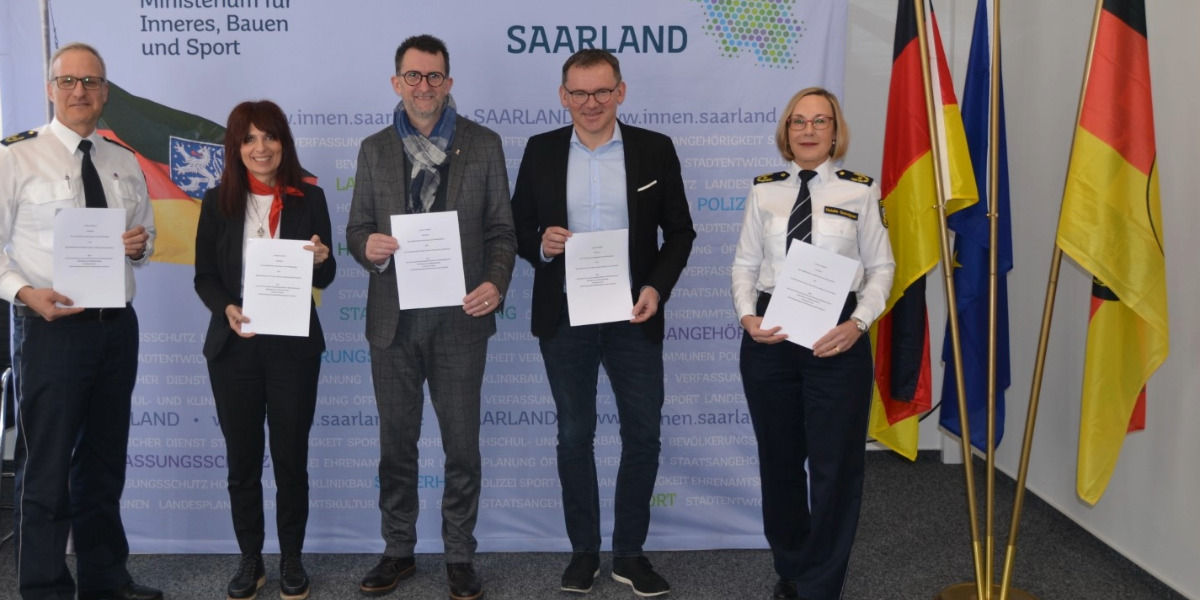 Drei Fahnen, drei Männer und zwei Frauen stehend. Sie halten die Vereinbarung über die Sicherheitspartnerschaft in der Hand.