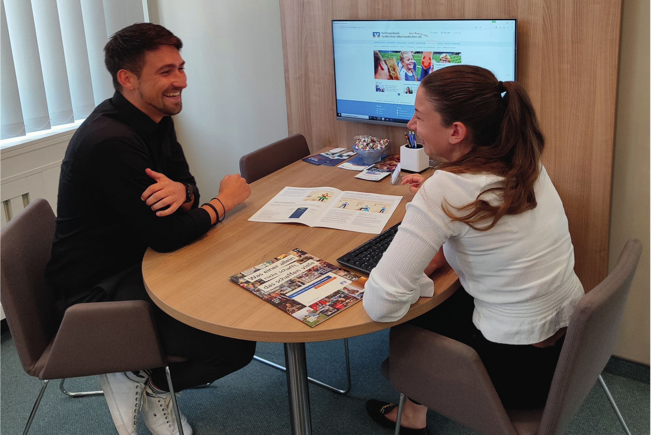 Zwei junge Erwachsene sitzen an einem Beratertisch und sprechen miteinander. Sie lachen. Dahinter ist ein Bildschirm zu sehen.
