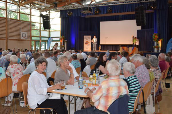 Foto der Hauptversammlung von 2019, auf dem viele Menschen an Tischen sitzen und eine Bühne im Hintergrund zu sehen ist