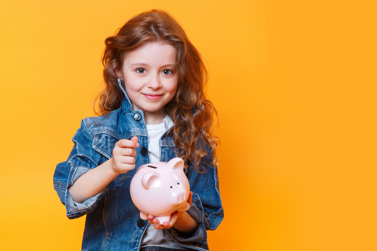 Kind mit langen, lockigen Haaren in Jeansjacke hält ein rosafarbenes Sparschwein vor orangefarbenem Hintergrund.