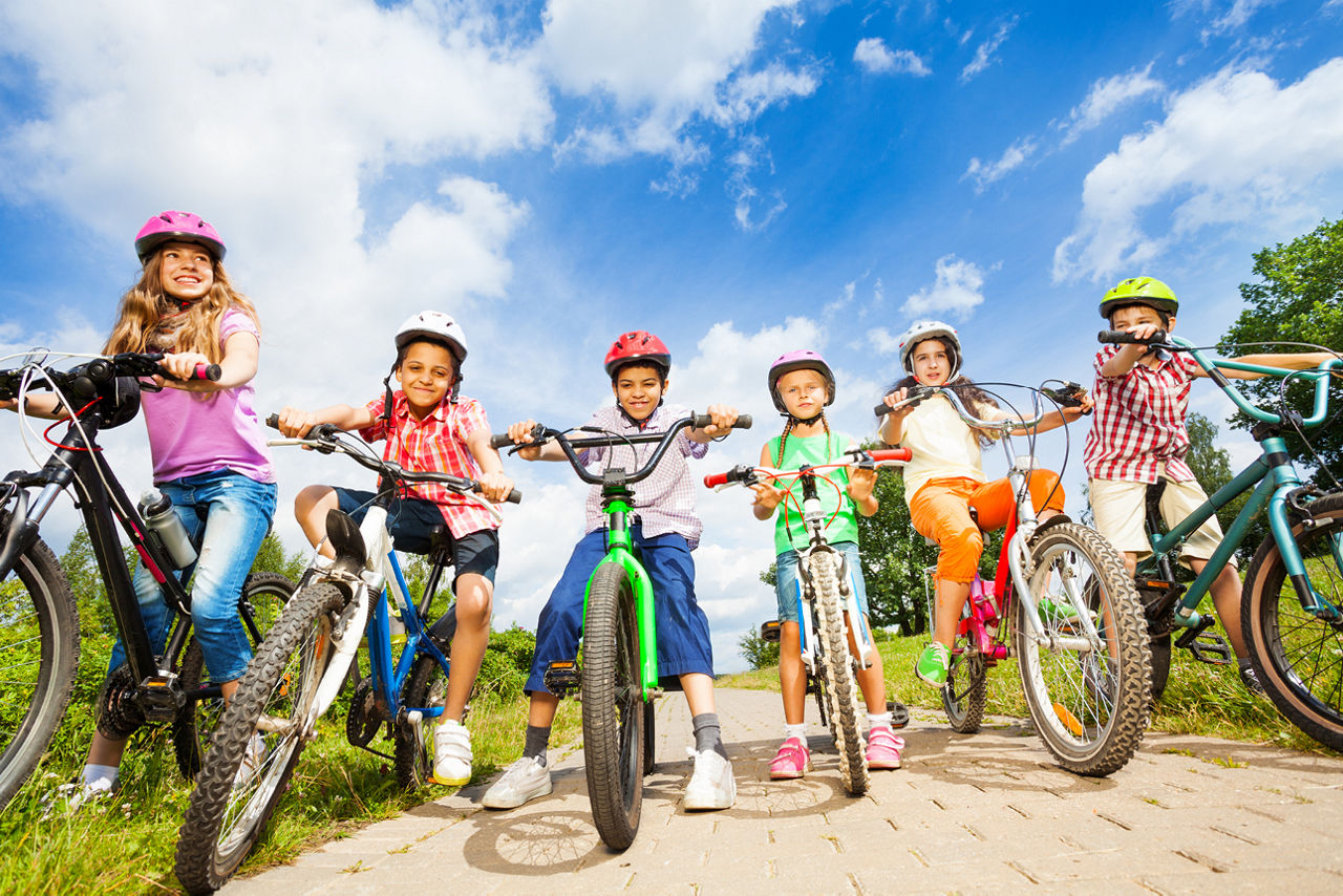 Sechs Kinder mit Fahrradhelmen sitzen auf Fahrrädern auf einem gepflasterten Weg unter blauem Himmel.