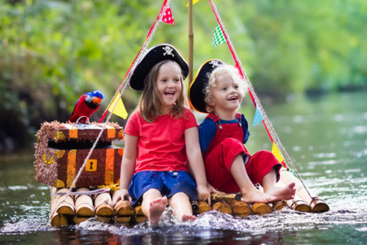 Zwei Kinder mit Piratenhüten sitzen barfuß auf einem Floß aus Bambusstäben, das mit bunten Wimpeln und einer Schatztruhe mit 
