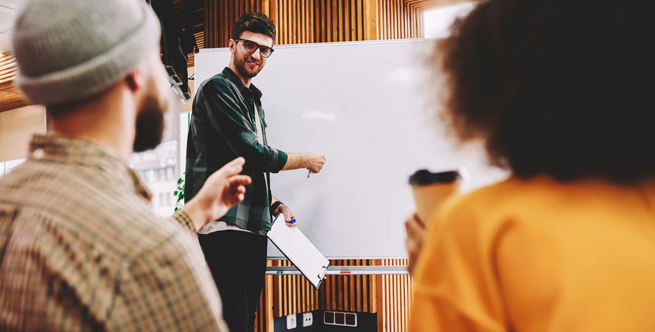 Mann mit Brille stehend vor Whiteboard und zwei Personen sitzend im Vordergrund