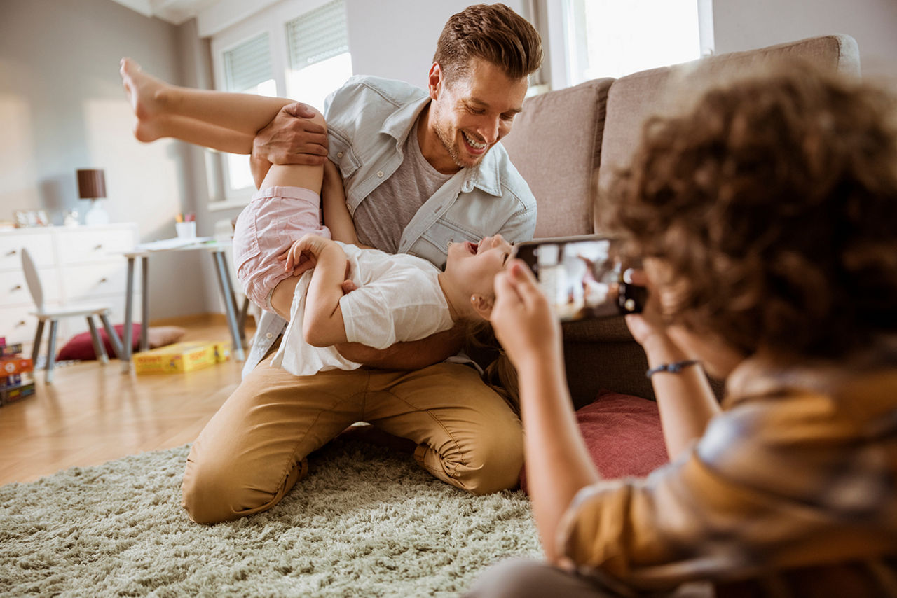 Vater und Kind albern im Wohnzimmer und werden dabei von dritter Person mit Handy fotografiert