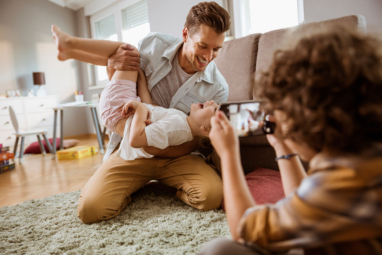 Vater und Kind albern im Wohnzimmer und werden dabei von dritter Person mit Handy fotografiert
