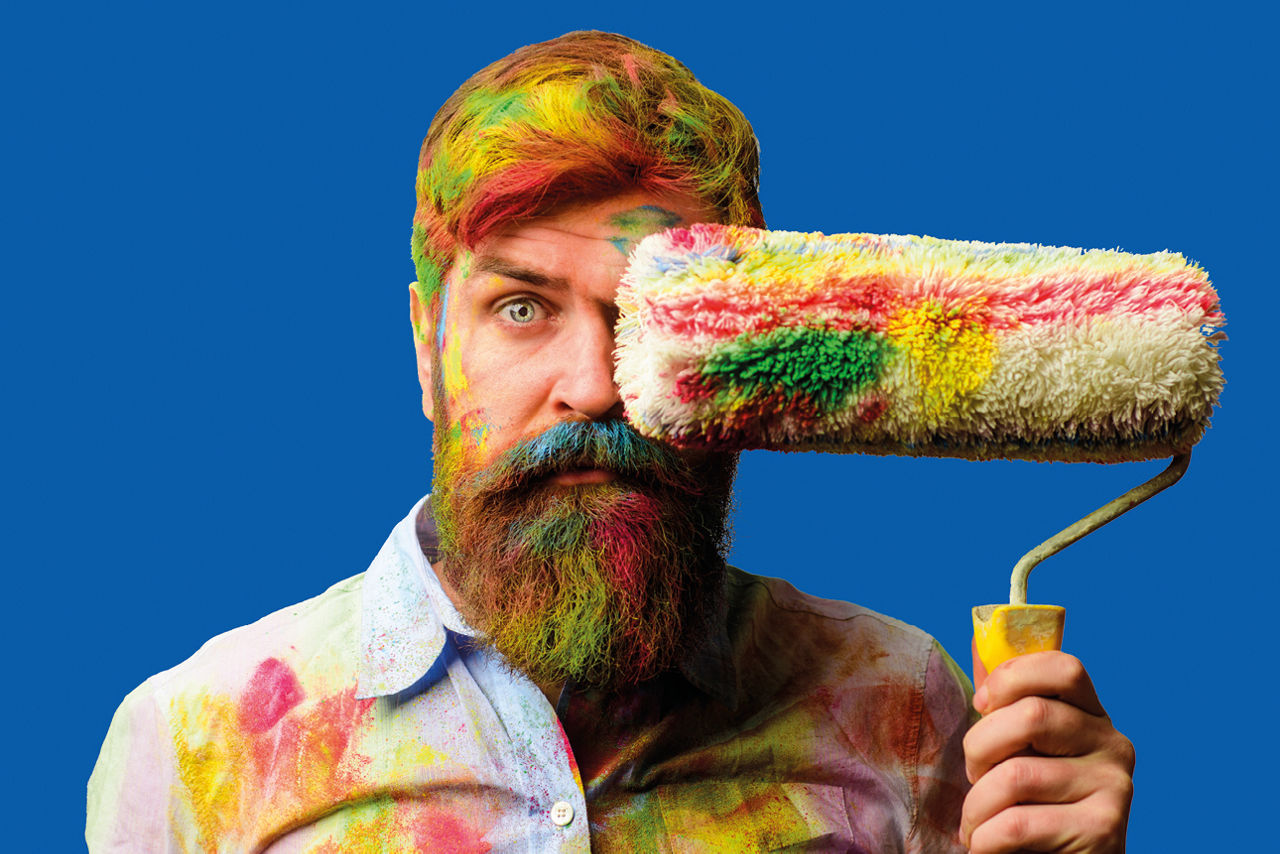 Mann steht vor einem blauen Hintergrund, hält eine Farbrolle in der Hand und ist bunt mit Farbe verschmiert