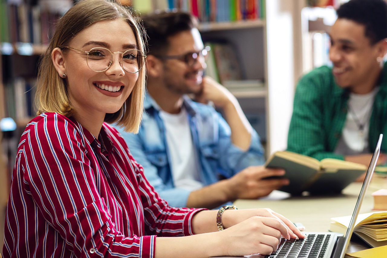   Drei Studierende lernen in der Bibliothek, eine Studentin sitzt vor ihrem Laptop und schaut in die Kamera.