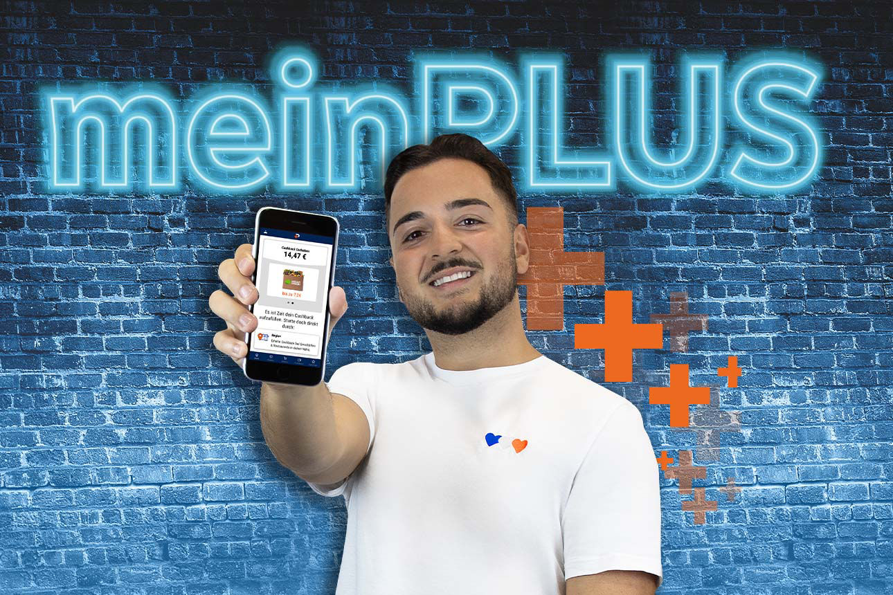 Junger Mann hält Smartphone mit geöffneter App hoch, im Hintergrund leuchtender Schriftzug ‚meinPLUS‘ auf blauer Ziegelwand.