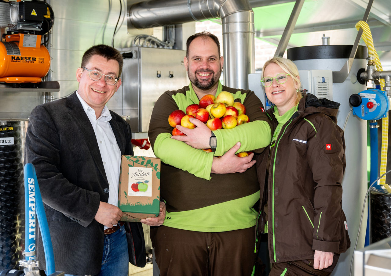 Ein Berater mit zwei Kunden vor der Saftpresse mit Äpfeln in der Hand