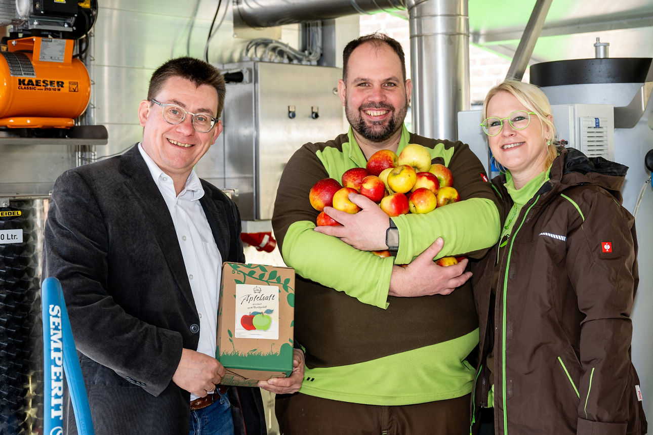 Ein Berater mit zwei Kunden vor der Saftpresse mit Äpfeln in der Hand