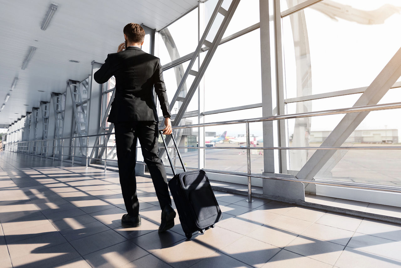Mann mit Handy am Ohr in einem Flughafen Gate zieht einen kleinen Handgepäckkoffer hinter sich her
