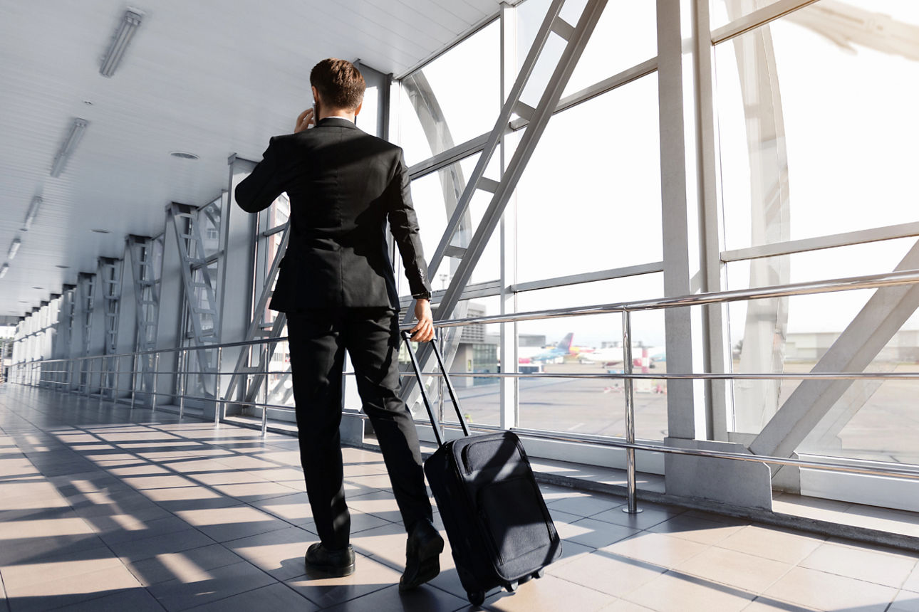 Mann mit Handy am Ohr in einem Flughafen Gate zieht einen kleinen Handgepäckkoffer hinter sich her