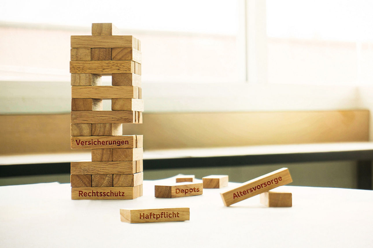 Turm aus Holzbausteinen mit einzelnen herausgezogenen Steinen, beschriftet mit Begriffen wie 'Versicherungen', 'Rechtsschutz', 'Depot', 'Haftpflicht' und 'Altersvorsorge'.