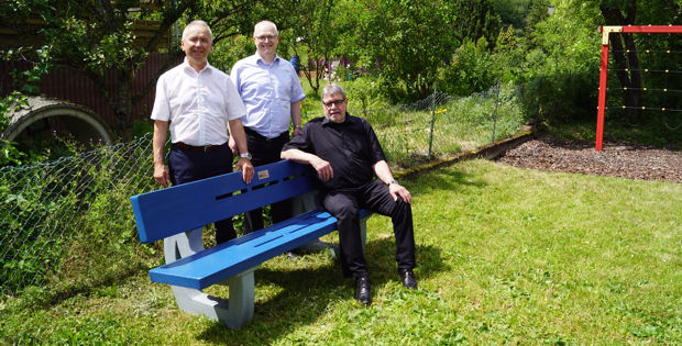 Drei Männer stehen und sitzen auf einer blauen Bank auf einer Wiese.