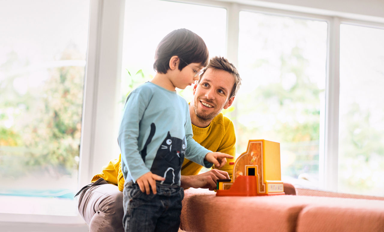Ein Mann und ein kleiner Junge spielen am Fenster gemeinsam mit einer Spielzeug-Kasse.